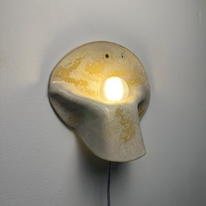 May include: A wall-mounted sconce featuring a curved, organic ceramic form with a textured yellow and white glaze. A single glowing spherical light bulb is positioned in the center of the fixture, which includes a small hole at the top for mounting.