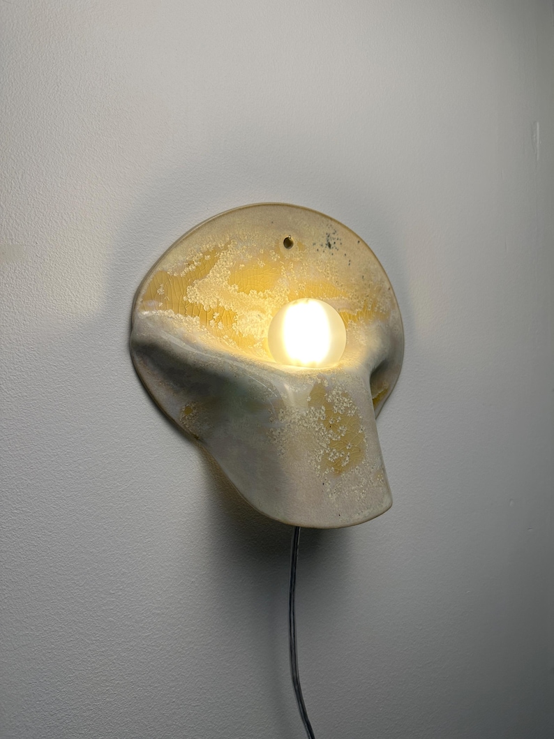 May include: A wall-mounted sconce featuring a curved, organic ceramic form with a textured yellow and white glaze. A single glowing spherical light bulb is positioned in the center of the fixture, which includes a small hole at the top for mounting.