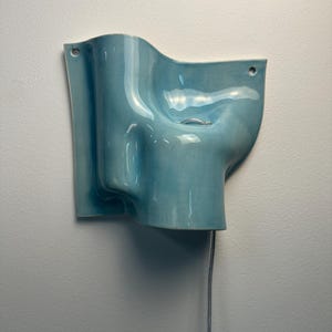 May include: A light blue ceramic wall sconce with a glossy finish and an undulating, organic shape. The fixture features two small circular holes for mounting and a single silver-toned metal ring detail. A thin, light-coloured electrical cord hangs down.