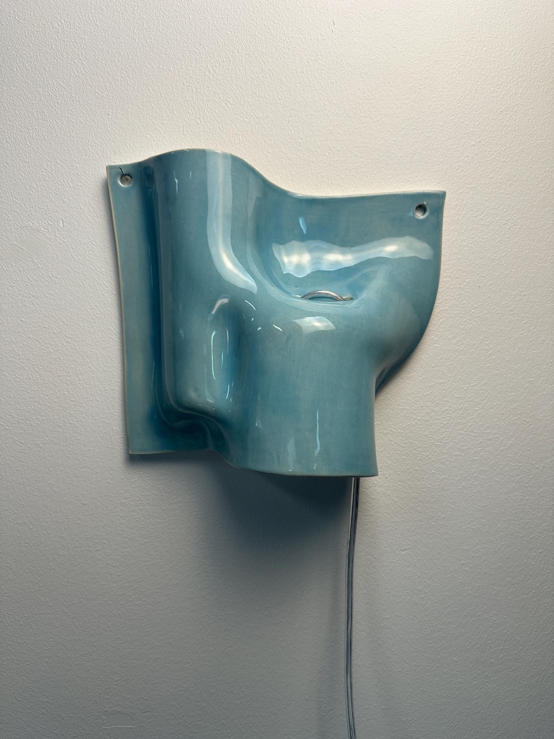 May include: A light blue ceramic wall sconce with a glossy finish and an undulating, organic shape. The fixture features two small circular holes for mounting and a single silver-toned metal ring detail. A thin, light-coloured electrical cord hangs down.