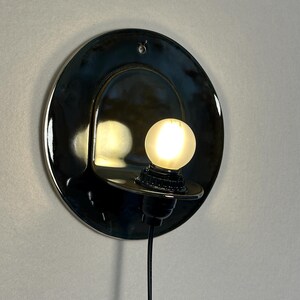 May include: A circular, glossy black ceramic wall sconce featuring a curved, protruding shelf that holds a small, glowing, frosted spherical light bulb. The fixture includes a black electrical cord extending downward from the base.