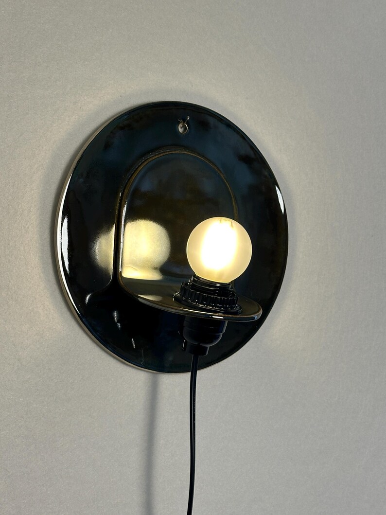 May include: A circular, glossy black ceramic wall sconce featuring a curved, protruding shelf that holds a small, glowing, frosted spherical light bulb. The fixture includes a black electrical cord extending downward from the base.