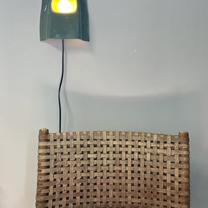 May include: A sage green ceramic wall light with a curved, rectangular form and a glowing bulb, mounted above a chair with a woven light brown seat back featuring a grid pattern and a black power cord with a switch hanging down.