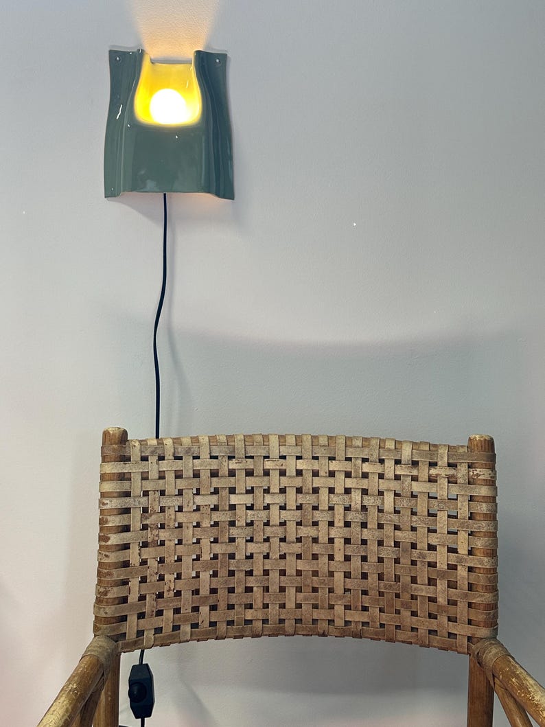 May include: A sage green ceramic wall light with a curved, rectangular form and a glowing bulb, mounted above a chair with a woven light brown seat back featuring a grid pattern and a black power cord with a switch hanging down.