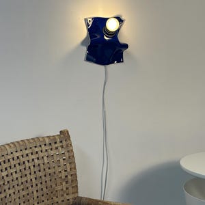 May include: A wall-mounted lamp featuring a glossy, deep blue, irregularly shaped ceramic base with a single exposed light bulb. The fixture is connected to a thin, light-colored electrical cord that hangs vertically down the wall.