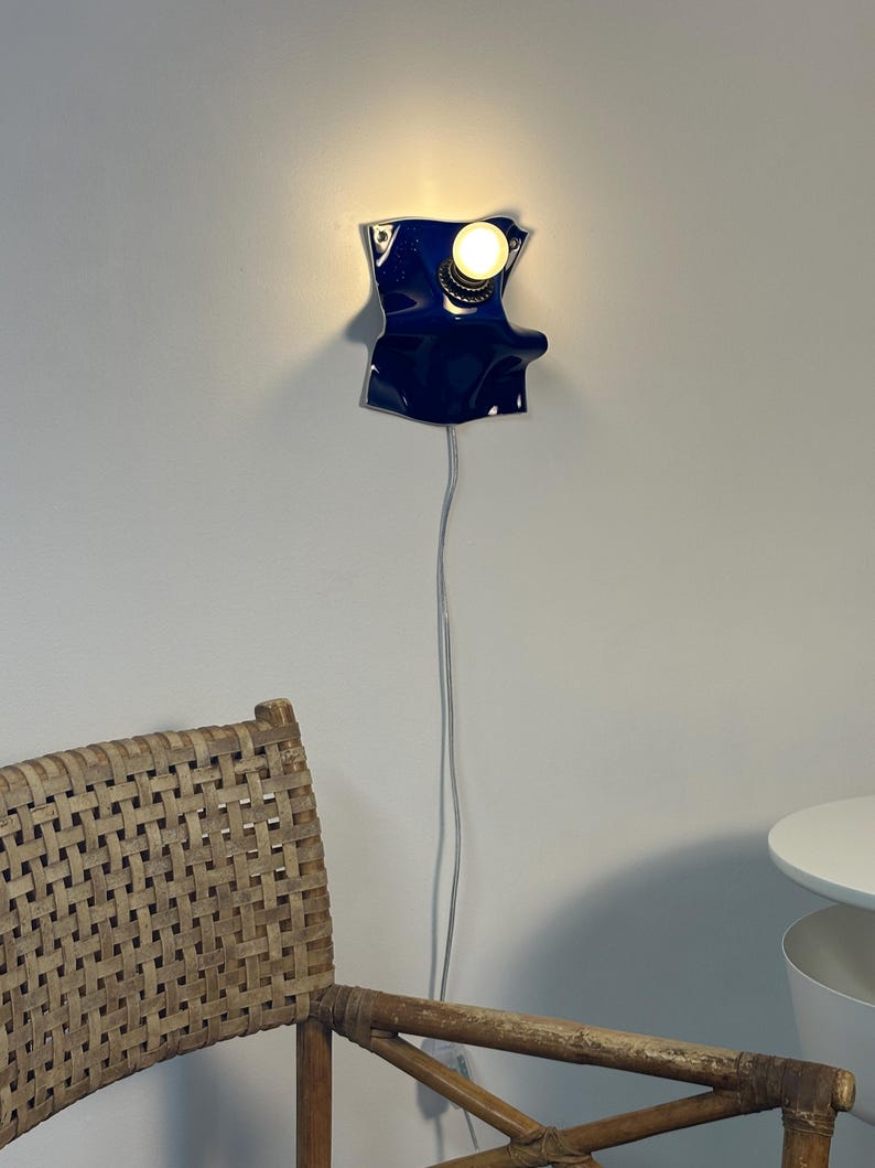 May include: A wall-mounted lamp featuring a glossy, deep blue, irregularly shaped ceramic base with a single exposed light bulb. The fixture is connected to a thin, light-colored electrical cord that hangs vertically down the wall.