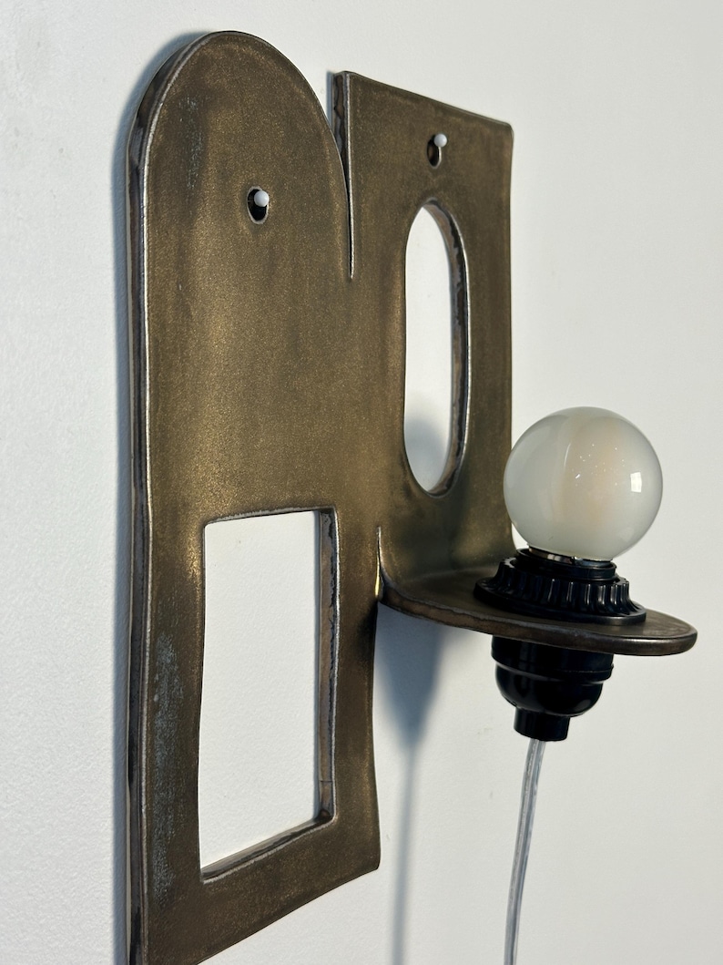 K&ouml;nnte beinhalten: Eine Wandleuchte mit einer flachen, bronzefarbenen Metallr&uuml;ckplatte, die oben abgerundet ist und einen rechteckigen Ausschnitt aufweist. Eine kleine, runde wei&szlig;e Gl&uuml;hbirne sitzt in einer schwarzen Fassung auf einer horizontalen Ablage.