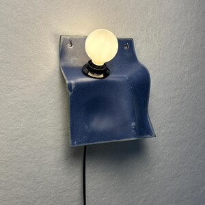May include: A wall-mounted lamp featuring a curved, blue ceramic base with a glossy finish. The fixture includes a black socket holding a single, round, glowing white light bulb. A thin black power cord extends downward from the base of the lamp.