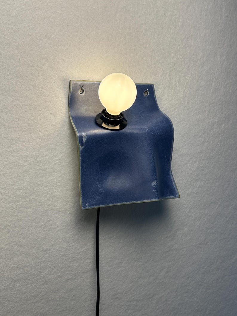 May include: A wall-mounted lamp featuring a curved, blue ceramic base with a glossy finish. The fixture includes a black socket holding a single, round, glowing white light bulb. A thin black power cord extends downward from the base of the lamp.