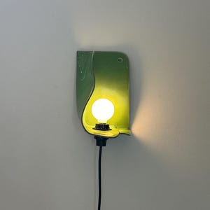 Pu&ograve; includere: Una lampada da parete con un paralume in ceramica curva di colore verde chiaro lucido. Una singola lampadina bianca rotonda e luminosa &egrave; centrata all'interno del paralume, fissata a un portalampada nero con un cavo nero che scende.
