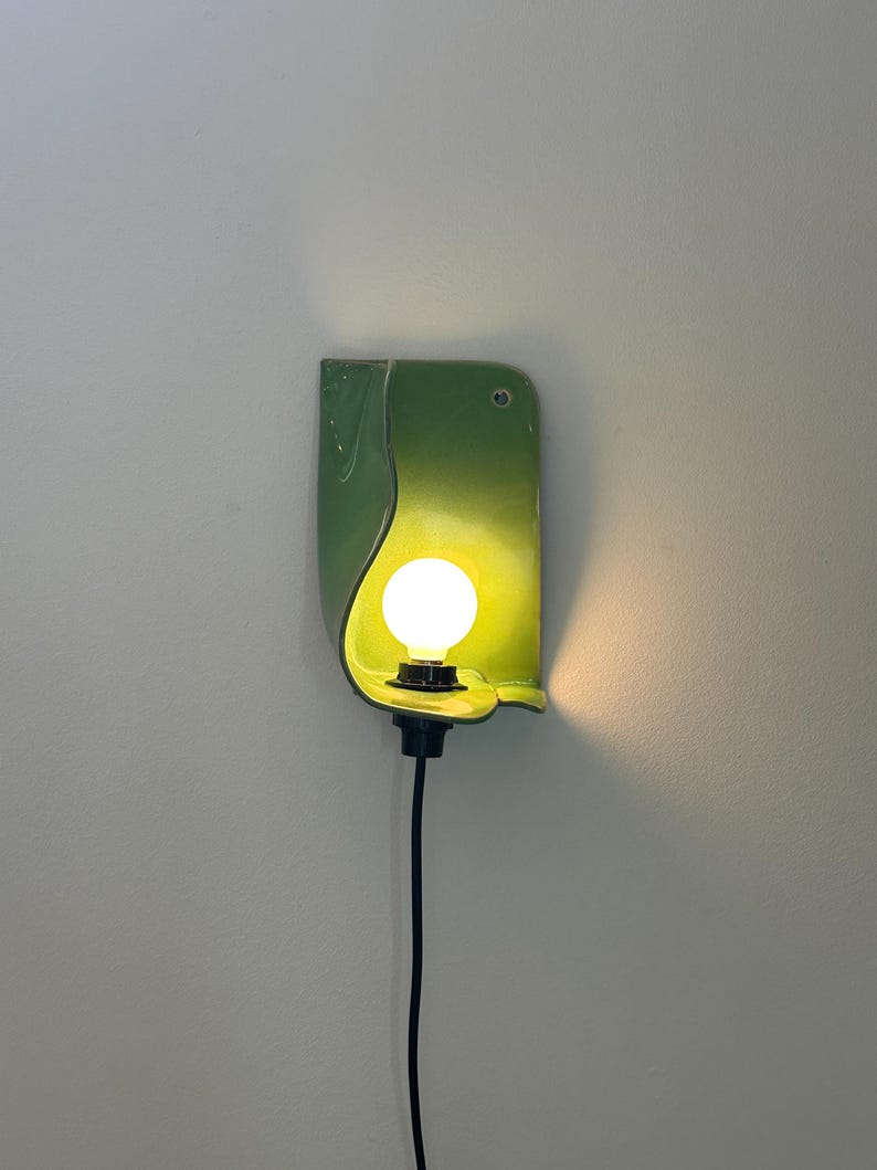 Pu&ograve; includere: Una lampada da parete con un paralume in ceramica curva di colore verde chiaro lucido. Una singola lampadina bianca rotonda e luminosa &egrave; centrata all'interno del paralume, fissata a un portalampada nero con un cavo nero che scende.