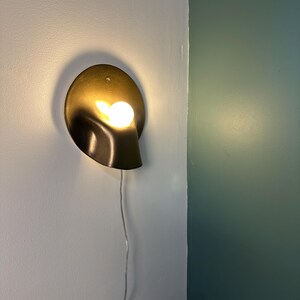 May include: A wall-mounted sconce featuring a circular, metallic gold-toned backplate with a curved, folded design. A single, round, warm-glowing light bulb is positioned within the fold of the fixture, which includes a thin, white power cord.