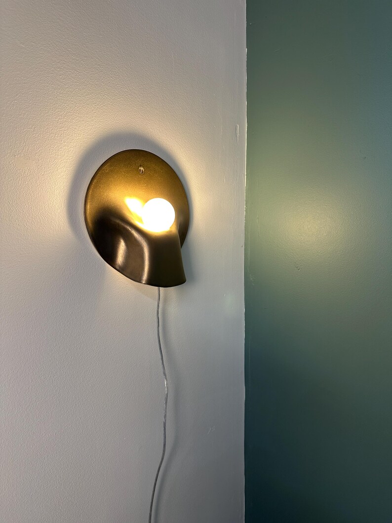 May include: A wall-mounted sconce featuring a circular, metallic gold-toned backplate with a curved, folded design. A single, round, warm-glowing light bulb is positioned within the fold of the fixture, which includes a thin, white power cord.