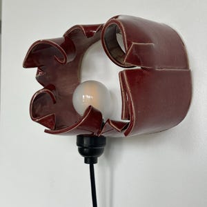May include: A burgundy ceramic wall sconce with a unique, curved design. The sconce features a light bulb and a black cord. The fixture is mounted on a white wall. The overall design is modern and artistic.