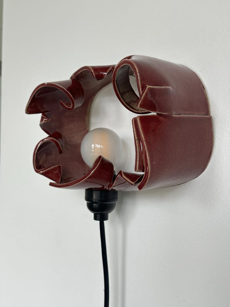 May include: A burgundy ceramic wall sconce with a unique, curved design. The sconce features a light bulb and a black cord. The fixture is mounted on a white wall. The overall design is modern and artistic.