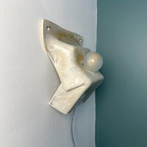May include: A wall-mounted ceramic sconce featuring an angular, folded design with a glossy, cream-colored glaze and subtle golden-yellow marbling. The fixture includes a single round, frosted glass light bulb protruding from the center.