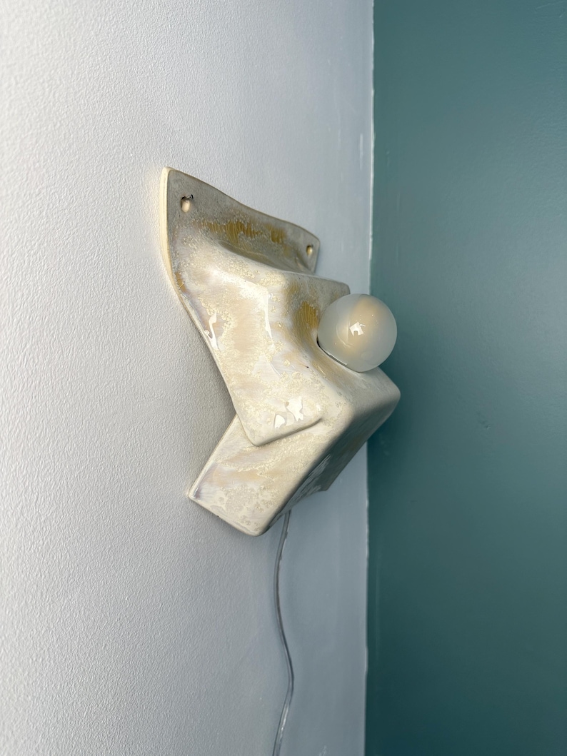 May include: A wall-mounted ceramic sconce featuring an angular, folded design with a glossy, cream-colored glaze and subtle golden-yellow marbling. The fixture includes a single round, frosted glass light bulb protruding from the center.