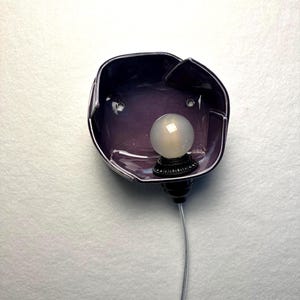 K&ouml;nnte beinhalten: Eine Wandleuchte mit einem tiefvioletten, unregelm&auml;&szlig;ig geformten Keramikschalen-Lampenschirm mit gl&auml;nzender Oberfl&auml;che. Eine kleine, runde, mattwei&szlig;e Gl&uuml;hbirne ist mittig in der Schale platziert, befestigt an einer schwarzen Fassung und einem d&uuml;nnen, transparenten Kabel.