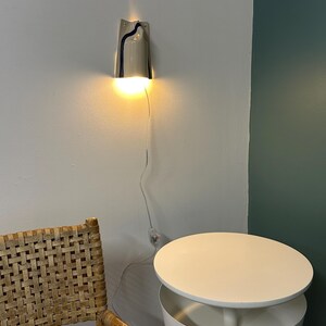 May include: A wall-mounted light fixture with a curved, light gray ceramic shade featuring a single dark blue vertical stripe. The lamp emits a warm, downward-facing glow and includes a thin, transparent power cord with an inline switch.