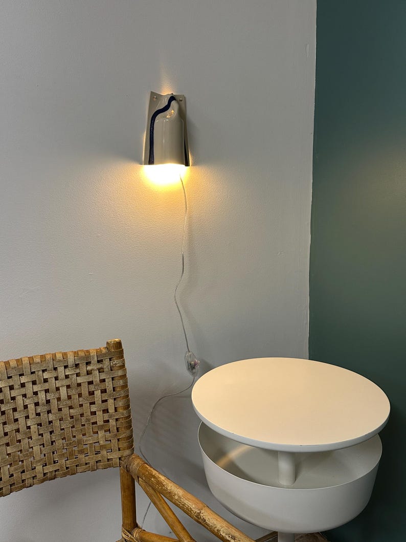 May include: A wall-mounted light fixture with a curved, light gray ceramic shade featuring a single dark blue vertical stripe. The lamp emits a warm, downward-facing glow and includes a thin, transparent power cord with an inline switch.