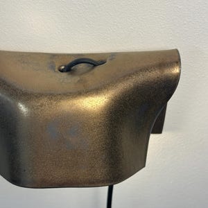 Pode incluir: Um candeeiro de parede com um abajur curvo em tom de bronze met&aacute;lico com um acabamento texturizado e salpicado. Um cabo el&eacute;trico preto sai do centro superior da lumin&aacute;ria, que est&aacute; montada numa superf&iacute;cie de parede lisa e clara.