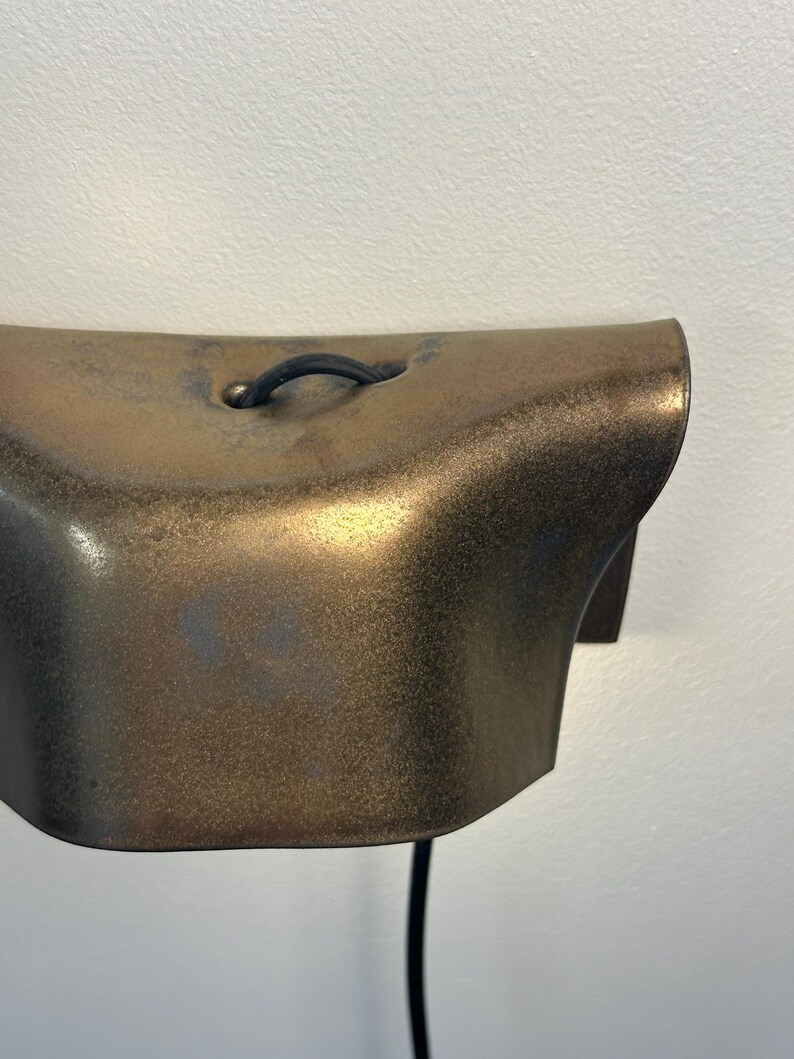 Pode incluir: Um candeeiro de parede com um abajur curvo em tom de bronze met&aacute;lico com um acabamento texturizado e salpicado. Um cabo el&eacute;trico preto sai do centro superior da lumin&aacute;ria, que est&aacute; montada numa superf&iacute;cie de parede lisa e clara.