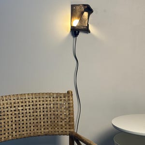 May include: A wall-mounted sconce featuring a dark, angular, metallic housing that directs light from a single bulb. The fixture is connected to a thin black power cord that hangs vertically down the wall, providing illumination.