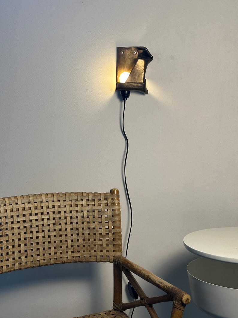 May include: A wall-mounted sconce featuring a dark, angular, metallic housing that directs light from a single bulb. The fixture is connected to a thin black power cord that hangs vertically down the wall, providing illumination.