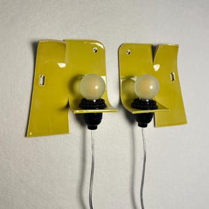 May include: A pair of wall-mounted light fixtures featuring glossy chartreuse ceramic backplates with irregular, folded edges. Each fixture includes a black socket holding a round, frosted glass light bulb and a thin, clear electrical cord.