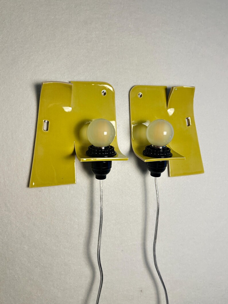May include: A pair of wall-mounted light fixtures featuring glossy chartreuse ceramic backplates with irregular, folded edges. Each fixture includes a black socket holding a round, frosted glass light bulb and a thin, clear electrical cord.
