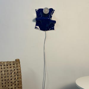 May include: A wall-mounted lamp featuring a glossy, deep blue, wavy ceramic or glass shade with a single exposed white light bulb. A thin, clear electrical cord hangs vertically from the base of the fixture toward the floor.