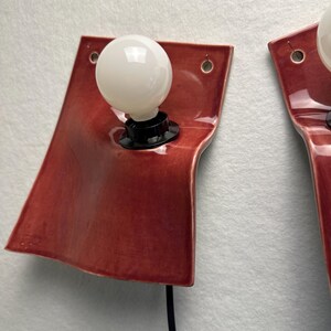 Puede incluir: Un aplique de pared con una placa de cer&aacute;mica roja oscura brillante de forma curva y org&aacute;nica. Una bombilla redonda de cristal blanco se inserta en un casquillo negro sobre la cer&aacute;mica. Se observan dos peque&ntilde;os orificios en las esquinas.