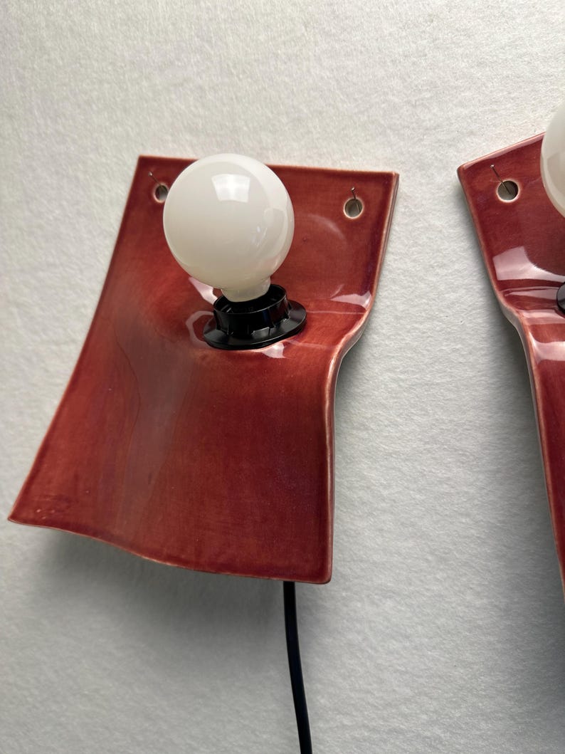 Puede incluir: Un aplique de pared con una placa de cer&aacute;mica roja oscura brillante de forma curva y org&aacute;nica. Una bombilla redonda de cristal blanco se inserta en un casquillo negro sobre la cer&aacute;mica. Se observan dos peque&ntilde;os orificios en las esquinas.