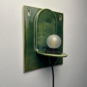 May include: A wall-mounted ceramic sconce featuring a glossy forest green finish. The fixture has a square backplate with two mounting holes and a central arched shelf holding a small, round, frosted white light bulb in a black socket.
