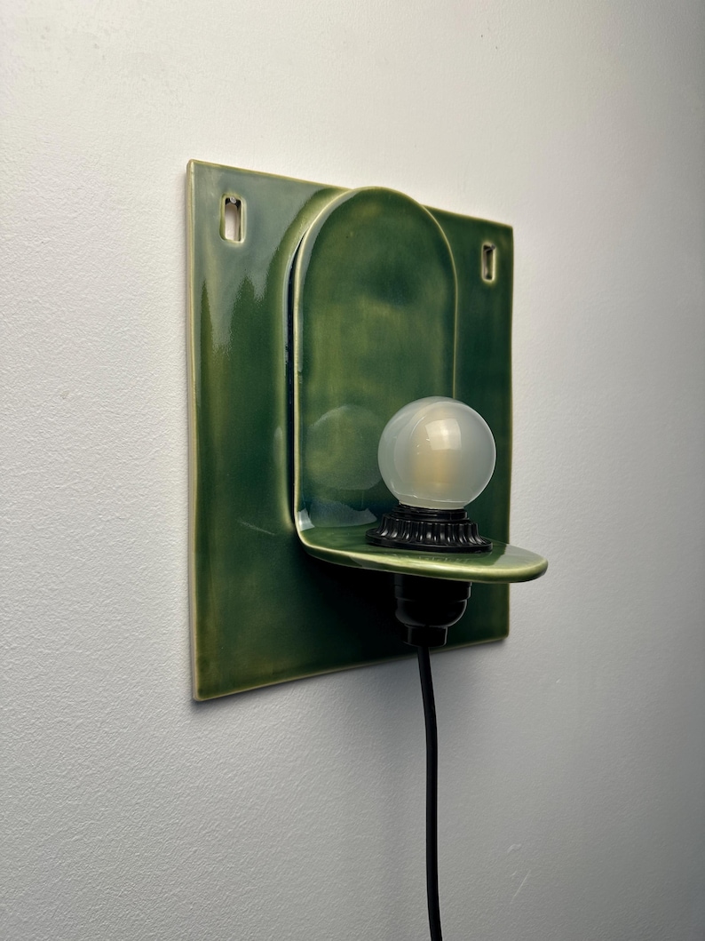 May include: A wall-mounted ceramic sconce featuring a glossy forest green finish. The fixture has a square backplate with two mounting holes and a central arched shelf holding a small, round, frosted white light bulb in a black socket.