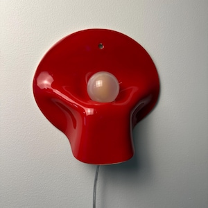 May include: A glossy red wall lamp with a curved, organic shape and a central circular opening. A single spherical white light bulb is positioned in the center of the red fixture. A thin gray electrical cord extends from the bottom of the lamp.