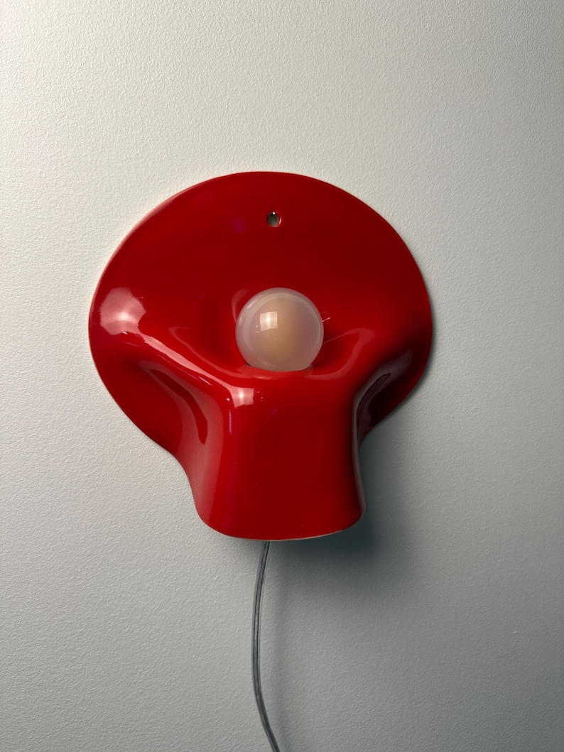 May include: A glossy red wall lamp with a curved, organic shape and a central circular opening. A single spherical white light bulb is positioned in the center of the red fixture. A thin gray electrical cord extends from the bottom of the lamp.