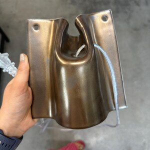 May include: A metallic bronze-colored wall-mounted fixture with a smooth, curved, and rectangular shape. It features a central U-shaped cutout at the top, two circular mounting holes at the upper corners, and a clear electrical cord threaded through.