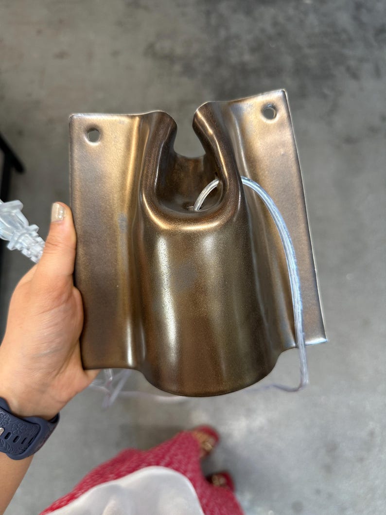 May include: A metallic bronze-colored wall-mounted fixture with a smooth, curved, and rectangular shape. It features a central U-shaped cutout at the top, two circular mounting holes at the upper corners, and a clear electrical cord threaded through.