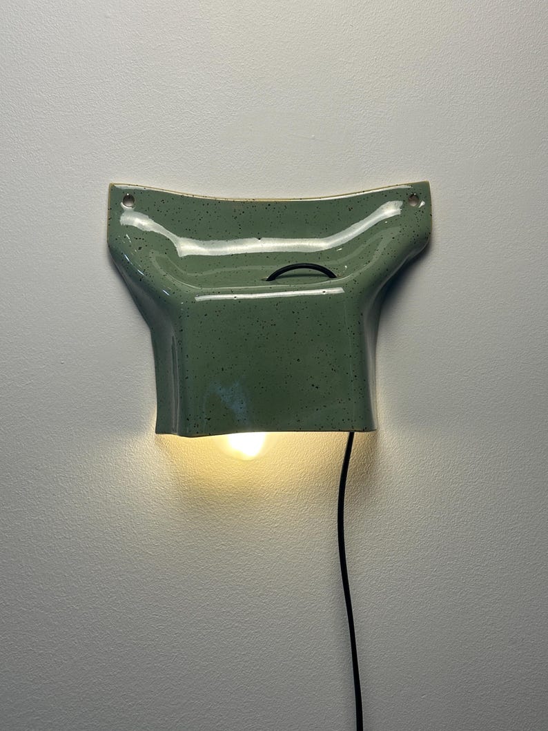 May include: A wall-mounted ceramic light fixture in a speckled sage green glaze. The piece features a flared, curved top section with two mounting holes and a downward-facing opening that emits a warm glow from an internal light bulb.