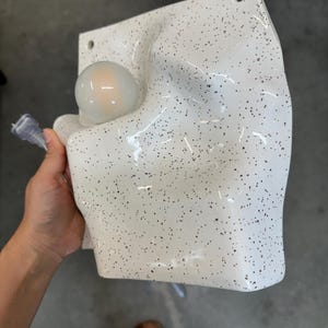 May include: A white wall-mounted lamp with a speckled black pattern and an irregular, molded surface. The fixture features a single round, frosted light bulb integrated into the left side and includes two circular mounting holes at the top corners.