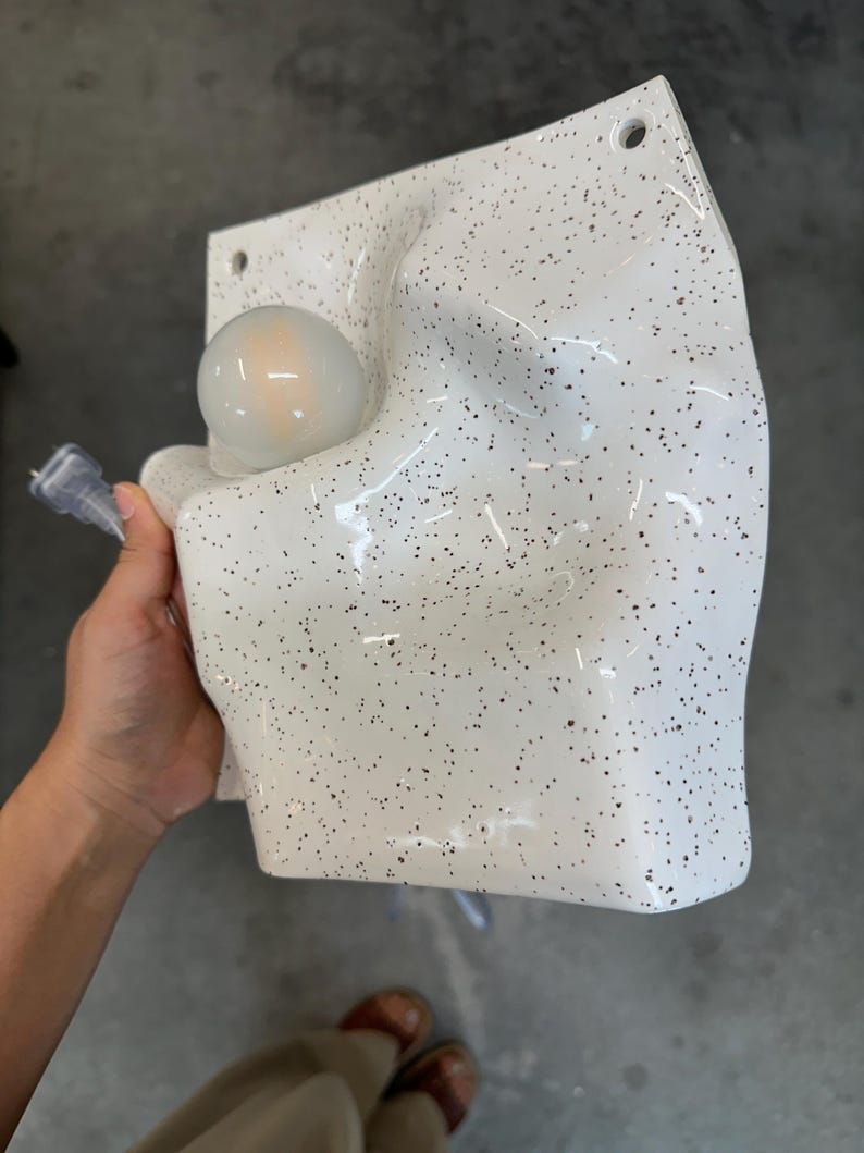 May include: A white wall-mounted lamp with a speckled black pattern and an irregular, molded surface. The fixture features a single round, frosted light bulb integrated into the left side and includes two circular mounting holes at the top corners.