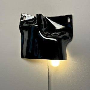 May include: A glossy black ceramic wall sconce featuring an irregular, draped, and folded surface texture. The fixture includes two rectangular mounting holes at the top corners and a single downward-facing light bulb emitting a warm glow.