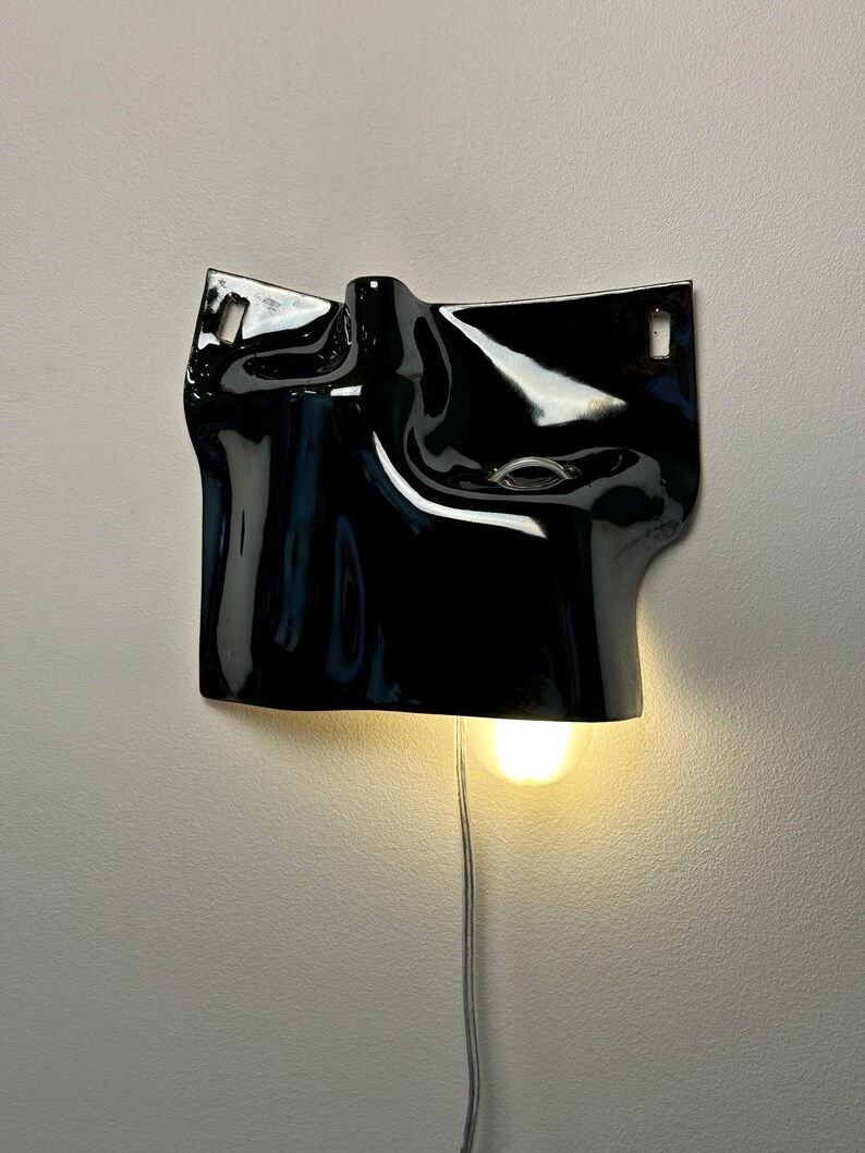 May include: A glossy black ceramic wall sconce featuring an irregular, draped, and folded surface texture. The fixture includes two rectangular mounting holes at the top corners and a single downward-facing light bulb emitting a warm glow.