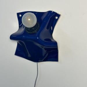 May include: A wall-mounted lamp featuring a glossy, deep blue ceramic base with an irregular, folded shape. The fixture includes a black socket holding a single, round, frosted white light bulb and a thin, light-colored electrical cord extending downward.