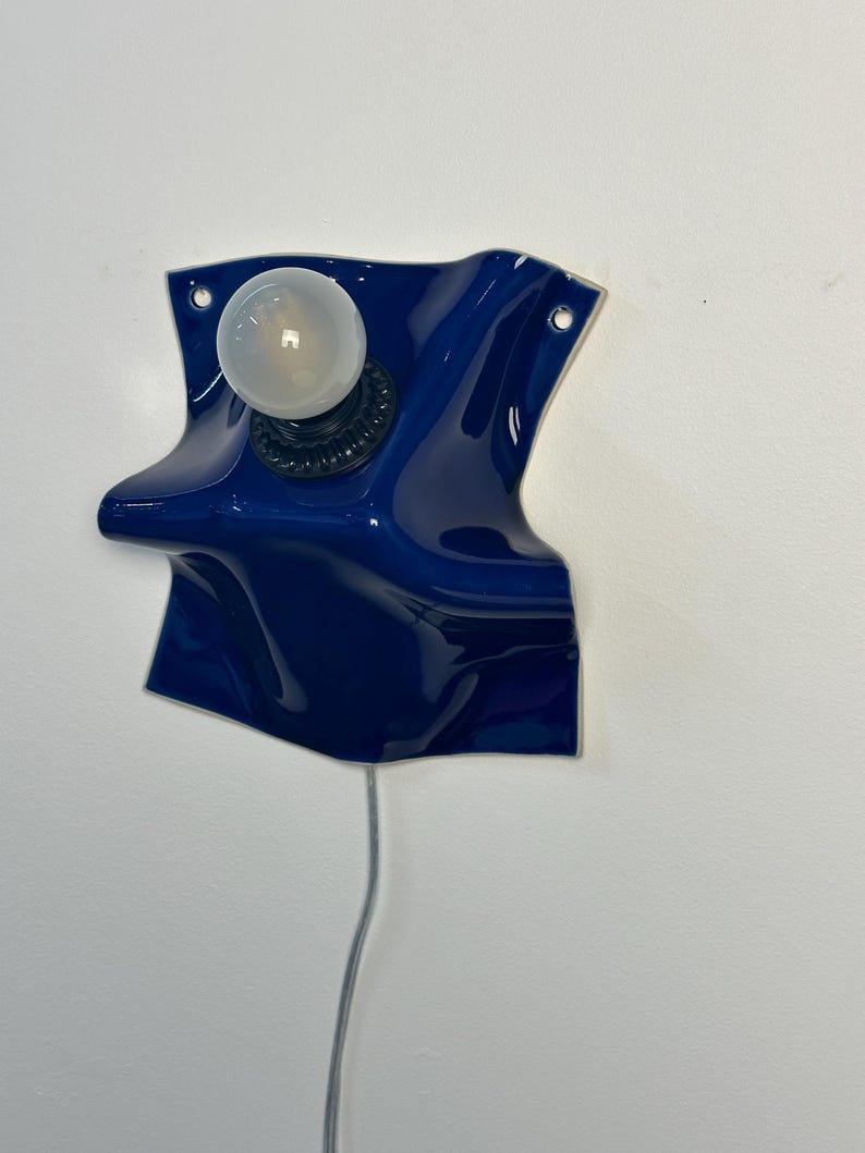 May include: A wall-mounted lamp featuring a glossy, deep blue ceramic base with an irregular, folded shape. The fixture includes a black socket holding a single, round, frosted white light bulb and a thin, light-colored electrical cord extending downward.