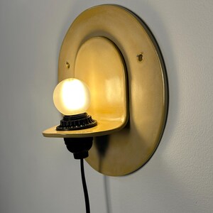 Pu&ograve; includere: Una lampada da parete con una piastra posteriore circolare in ceramica color giallo senape e un ripiano sporgente. Un portalampada nero sostiene una lampadina sferica smerigliata accesa e un cavo di alimentazione nero scende dalla base.