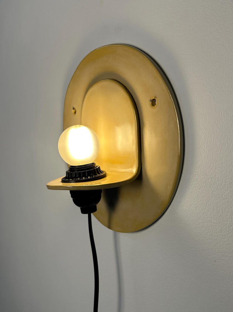 Pu&ograve; includere: Una lampada da parete con una piastra posteriore circolare in ceramica color giallo senape e un ripiano sporgente. Un portalampada nero sostiene una lampadina sferica smerigliata accesa e un cavo di alimentazione nero scende dalla base.