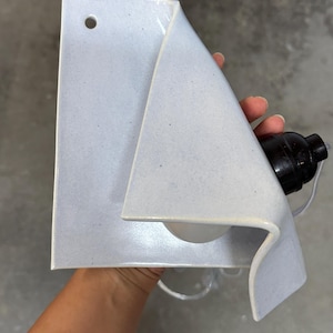 May include: A light blue ceramic wall sconce with a folded, sheet-like design. The fixture features a small circular hole at the top for mounting and includes a black electrical socket component attached to the side, designed to hold a light bulb.