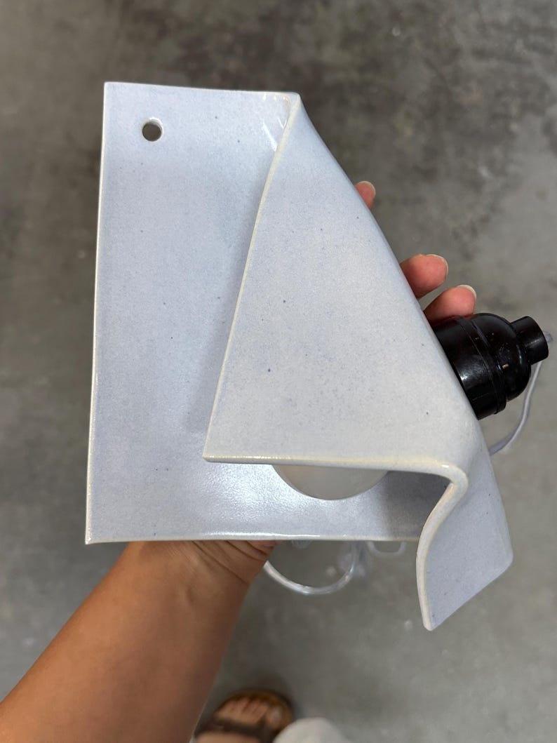 May include: A light blue ceramic wall sconce with a folded, sheet-like design. The fixture features a small circular hole at the top for mounting and includes a black electrical socket component attached to the side, designed to hold a light bulb.